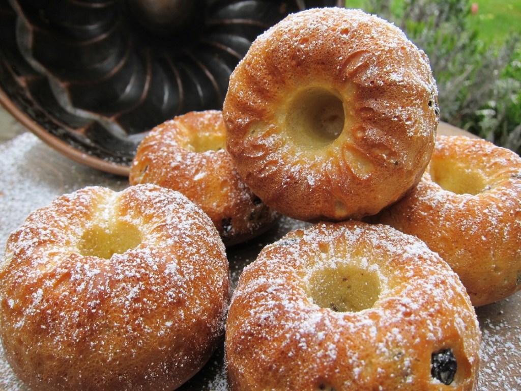 Sechs kleine runde Kuchen mit einer glatten Oberfläche und Puderzucker bestäubt, auf einem gedeckten Tisch.