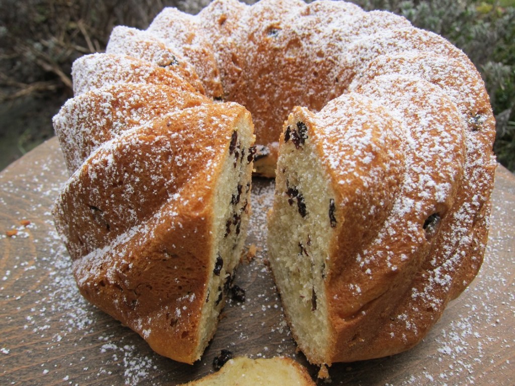 Ein Kuchen in einer Gugelhupfform, bestäubt mit Puderzucker und mit Rosinen gefüllt, auf einem Holzbrett serviert.