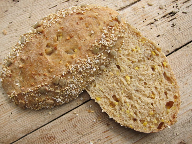 Ein rustikales Brot mit einer knusprigen Kruste, bestreut mit Saaten und Haferflocken, auf einem Holztisch in einer halbierten Ansicht.