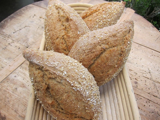 Eine Gruppe von vier rustikalen Brötchen, die mit Haferflocken bestreut sind, in einem Korb auf einer Holztischoberfläche.
