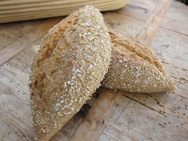 Zwei gebackene Brötchen aus Vollkornmehl, mit einer goldbraunen Kruste und bestreut mit Haferflocken, auf einem Holzuntergrund.