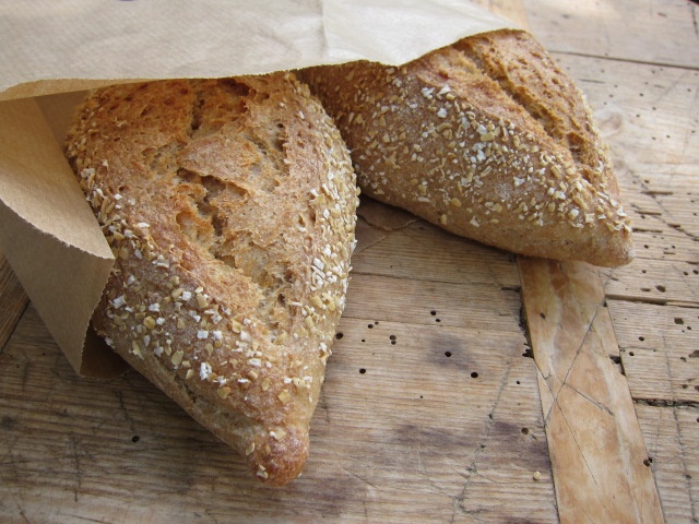 Zwei frisch gebackene Brötchen mit Haferflocken, in einer Papiertüte auf einem Holzuntergrund.