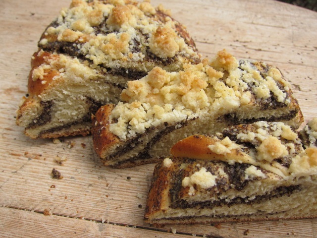 Ein aufgeschnittener Mohnzopf mit einer goldbraunen Kruste und zerbröseltem Topping auf der Oberfläche, auf einem Holzbrett präsentiert.
