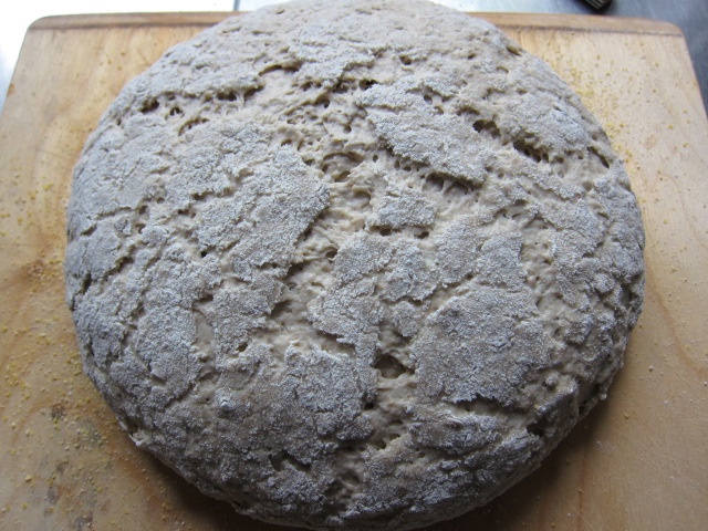 Ein runder Laib Brot mit einer rauen, mehlierten Oberfläche auf einem Holzbrett.