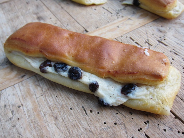 Ein längliches Gebäck, gefüllt mit einer süßen Creme und Rosinen, auf einem rustikalen Holzuntergrund.