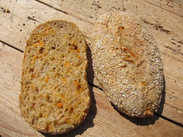 Ein frisch gebackenes Vollkornbrot mit einer goldenen Kruste und sichtbaren Haferflocken, auf einem Holztisch präsentiert. Eine Scheibe des Brotes, auf der die orangefarbenen Karottenstückchen sichtbar sind, liegt neben dem ganzen Brot.