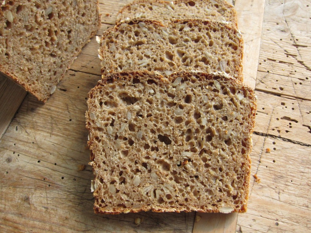 Scheiben von einem rustikalen Vollkornbrot, das auf einem Holzbrett liegt. Das Brot hat eine luftige Krume und ist mit Haferflocken und gepressten Körnern bestreut.