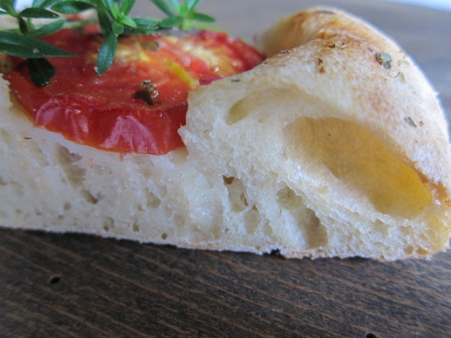 Nahaufnahme eines Stücks Ciabatta-Brot mit Tomatenscheiben und frischen Kräutern, auf einem Holzuntergrund.