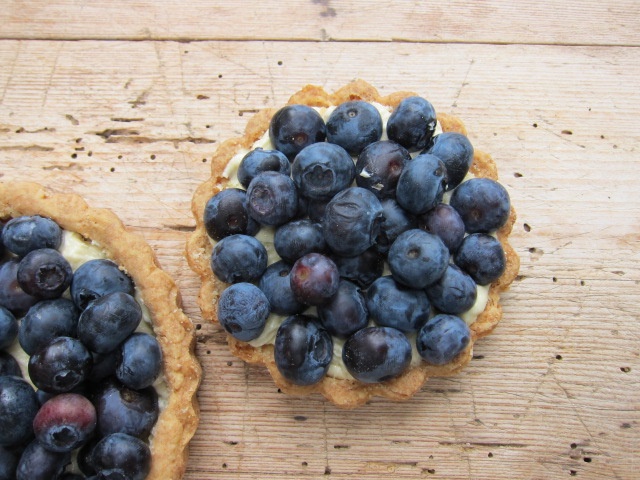 Zwei kleine Torten mit einer Füllung aus Vanillecreme und frischen Blaubeeren darauf, auf einem Holzuntergrund platziert.