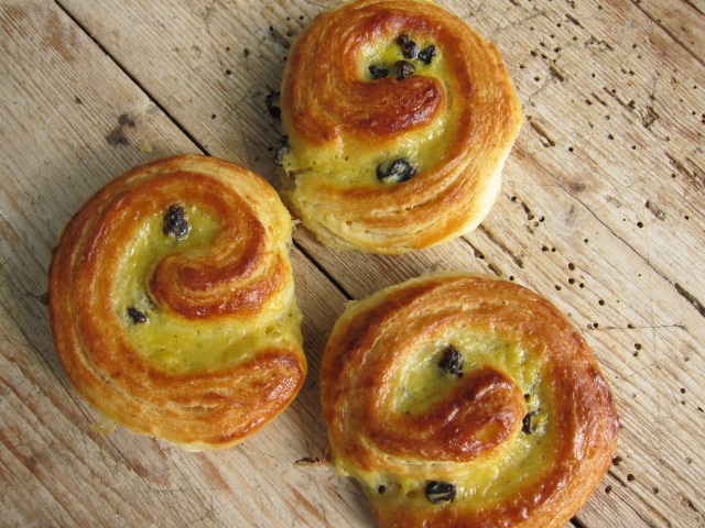 Drei süße Franzbrötchen mit einer goldbraunen, glänzenden Kruste und einem spiralförmigen Muster, auf einem rustikalen Holztisch.