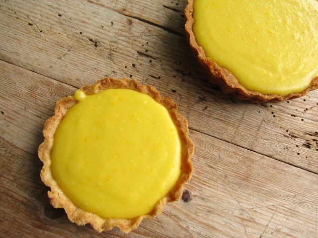 Zwei gelbe Zitronentörtchen in einem Mürbeteigboden, auf einem Holzuntergrund.