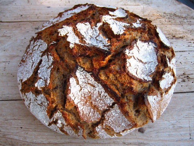 Rundes, hausgemachtes Brot mit einer rustikalen Kruste und einer goldbraunen Oberfläche, auf einem Holzbrett platziert.