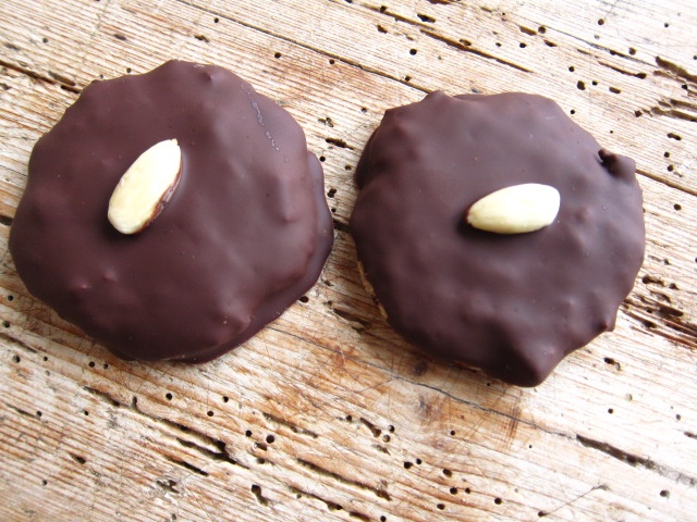 Zwei runde Kekse, überzogen mit dunkler Schokolade und jeweils einer Mandel obendrauf, auf einer rustikalen Holzoberfläche platziert.