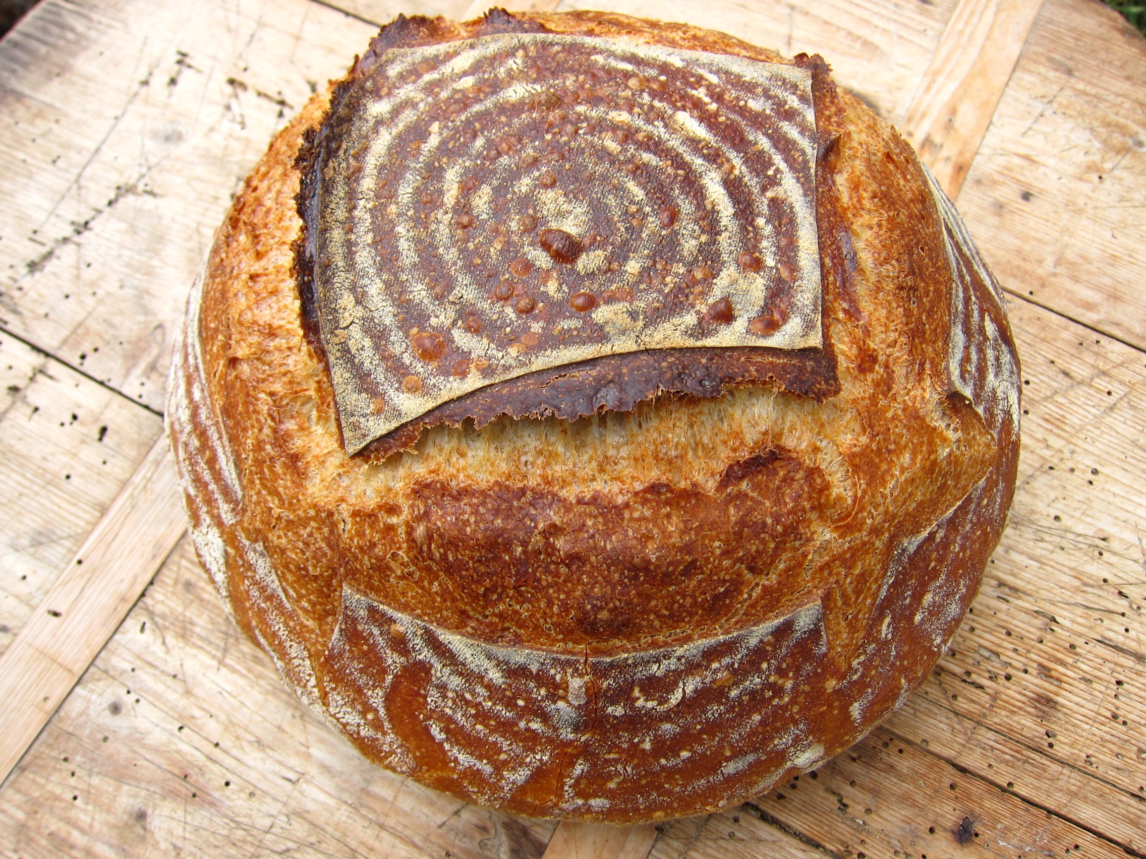 Ein rustikales Brot mit einer knusprigen Kruste und einem schönen Muster auf der Oberseite, auf einem Holzbrett platziert.