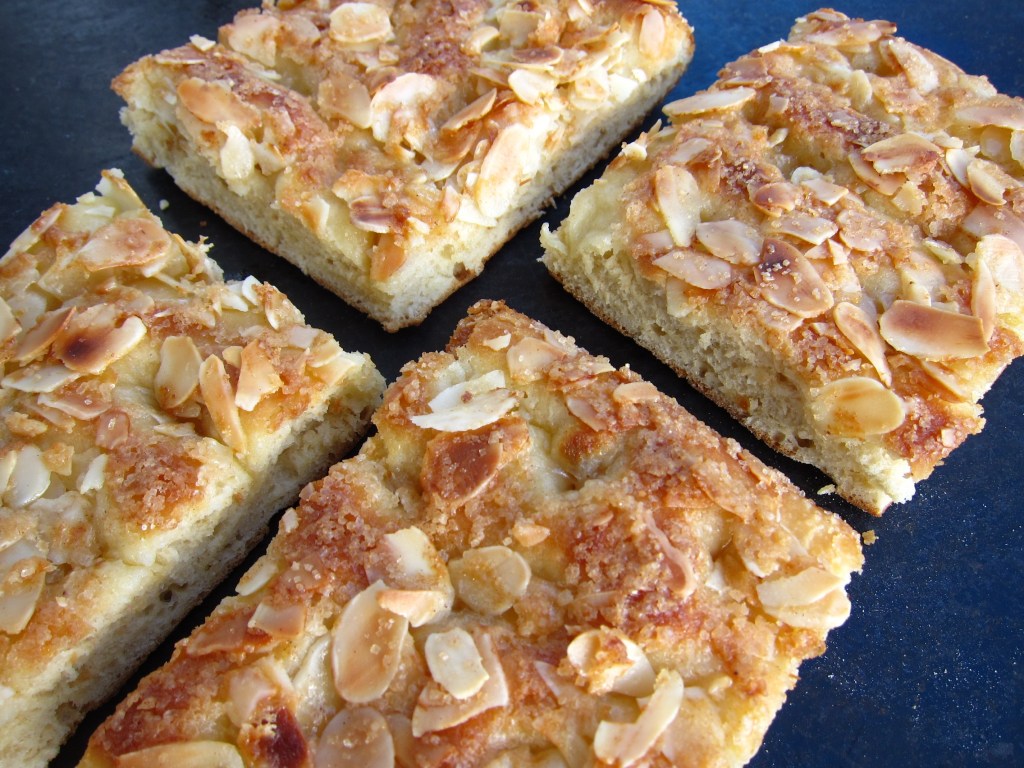 Ein gebackenes Dessert mit einer knusprigen, goldbraunen Oberseite und gehobelten Mandeln, in eckigen Stücken auf hellem Hintergrund.