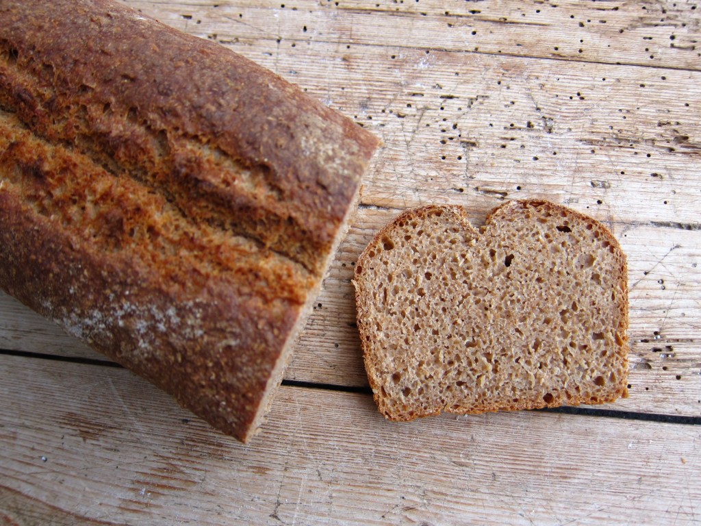 Ein Laib Roggenvollkornbrot mit einer geschnittenen Scheibe daneben auf einem Holzbrett.