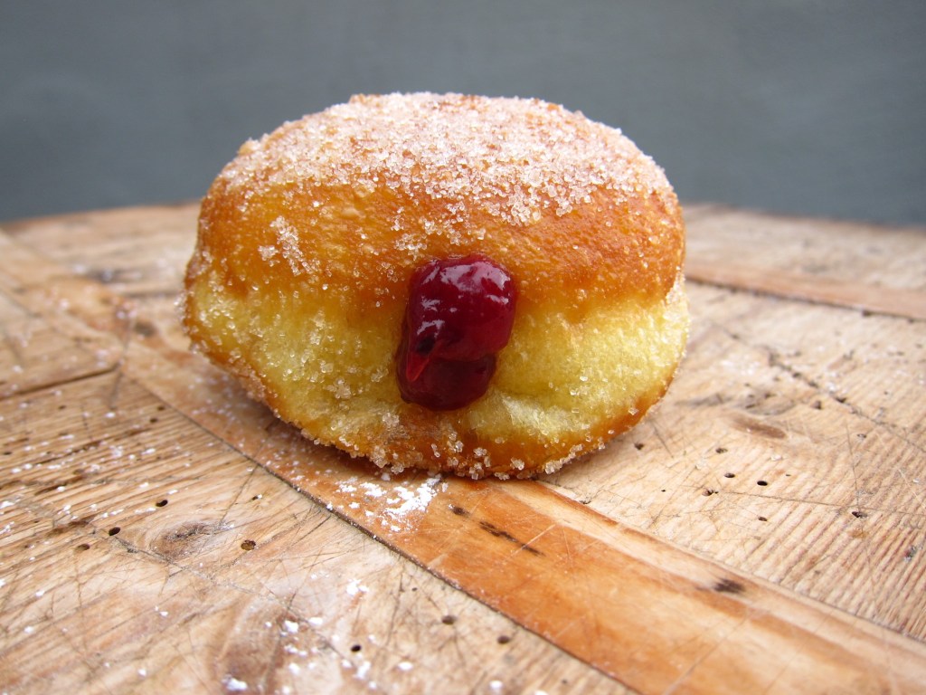 Ein gefüllter Berliner Pfannkuchen mit einer roten Marmelade, bestreut mit Zucker auf einem Holzuntergrund.