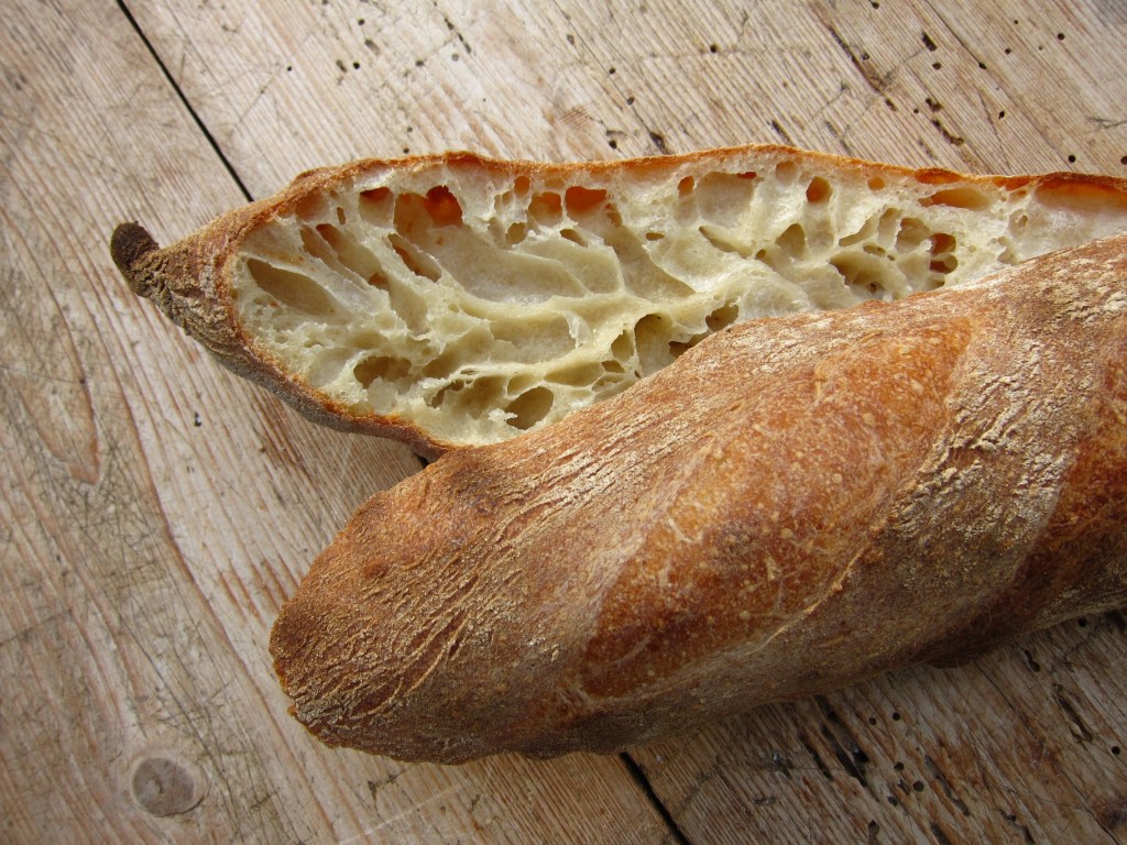 Ein aufgeschnittenes Baguette mit einer luftigen, offenen Krume und einer goldbraunen Kruste auf einem Holzuntergrund.