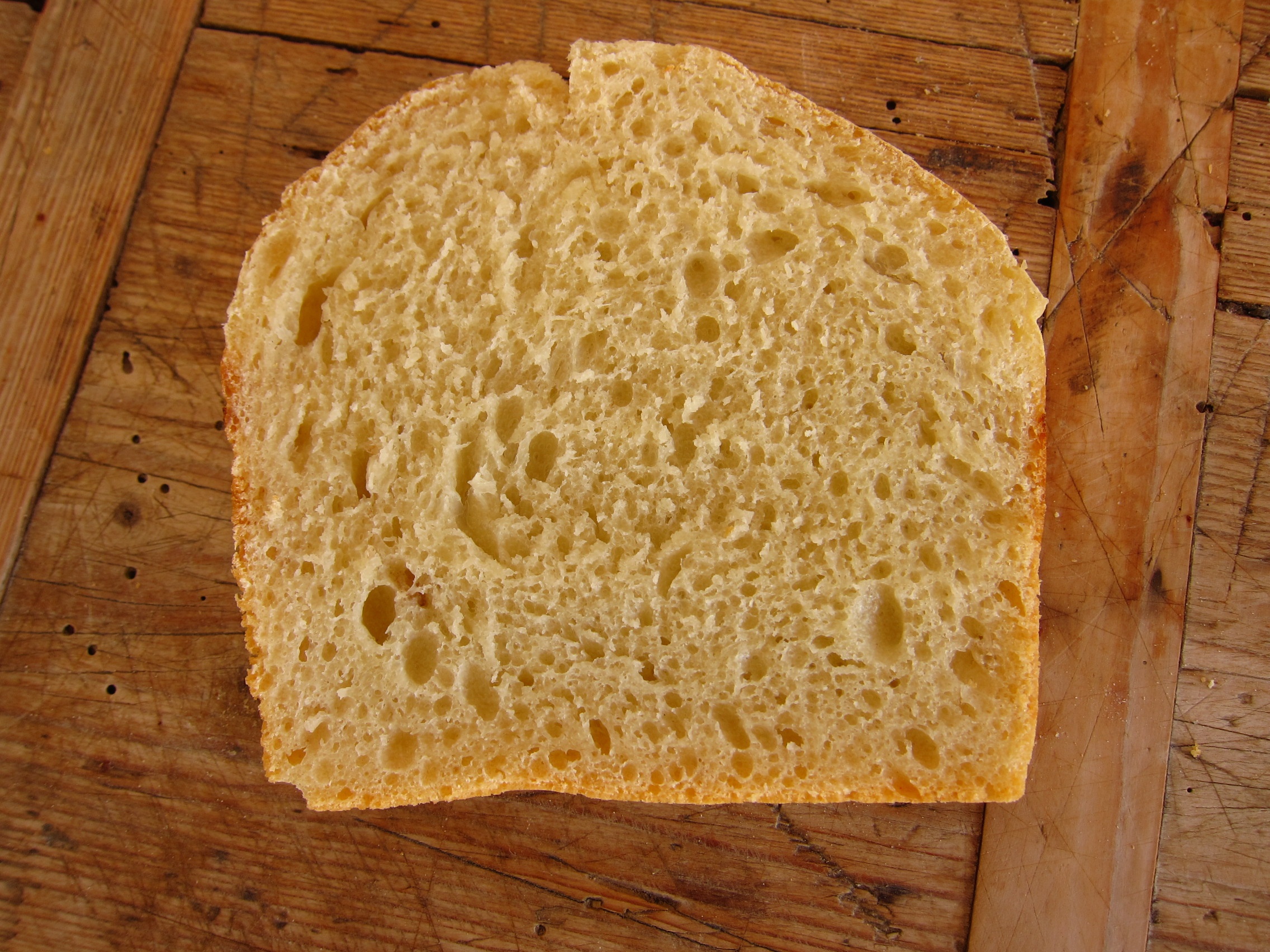 Eine aufgeschnittene Scheibe helles Brot mit einer luftigen Krume und goldbrauner Kruste, auf einem Holzbrett platziert.