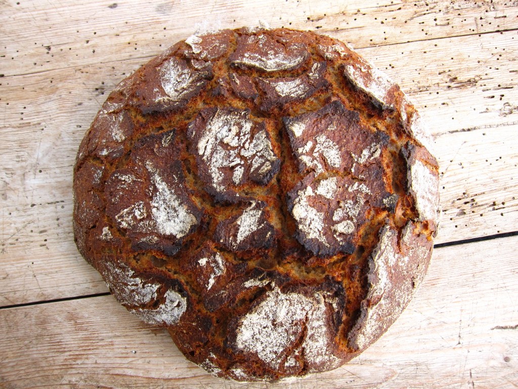 Ein rustikales, dunkelbraunes Brot mit einer knusprigen Kruste und rissiger Oberfläche, auf einem Holzbrett platziert.