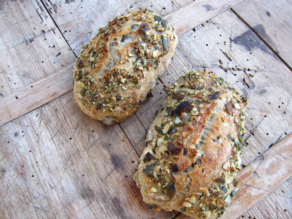 Zwei rustikale Brote mit einer knusprigen Kruste und einer Mischung aus verschiedenen Samen und Nüssen, auf einem Holztisch angeordnet.