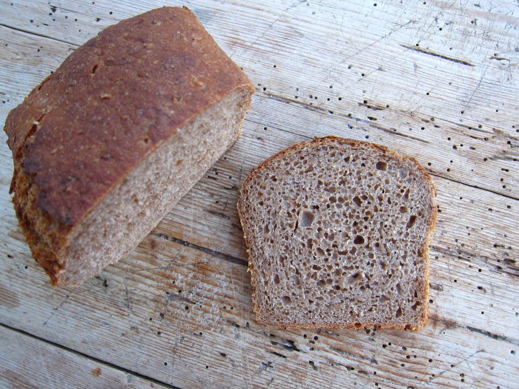Einlaib Roggenvollkornbrot auf einem Holztisch, mit einer Scheibe, die einen luftigen, kuchenartigen Innenraum zeigt.