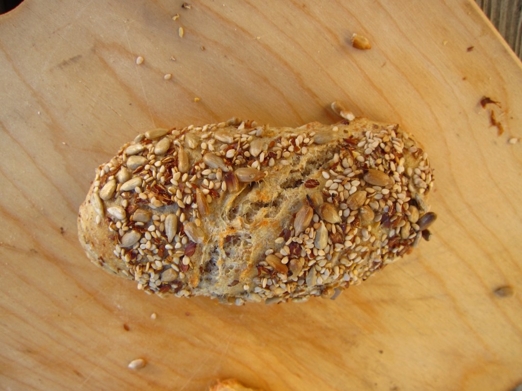 Ein unbestrichenes Brötchen mit Sesam und Sonnenblumenkernen auf der Oberseite, auf einer Holzplatte platziert.