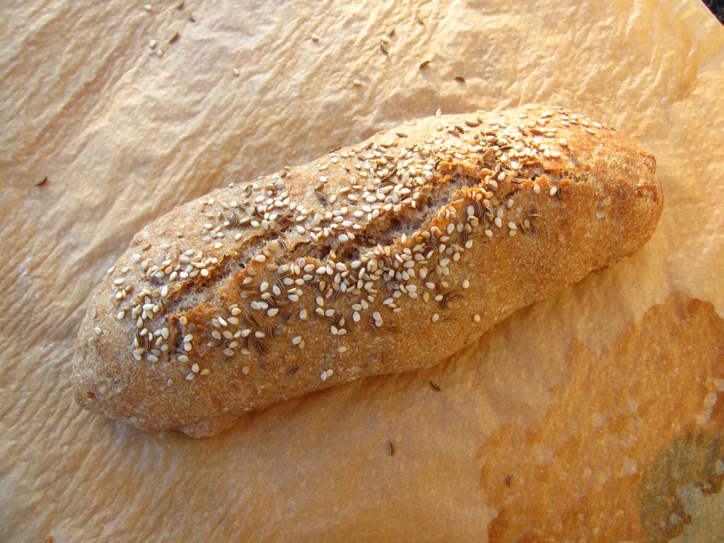 Ein frisches Dinkelbrötchen mit Sesam- und Kümmelsamen auf der Oberfläche, liegt auf einem Backpapier.