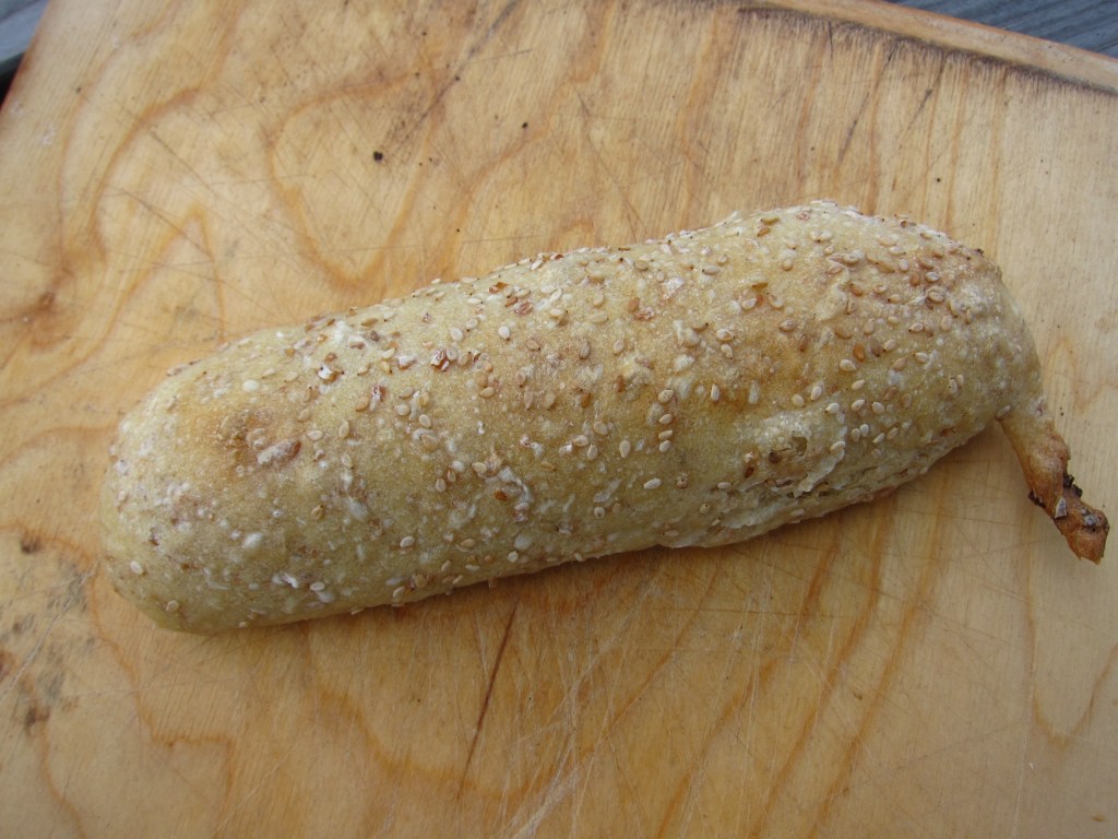Ein längliches Brötchen mit Sesamkruste auf einem Holzbrett.