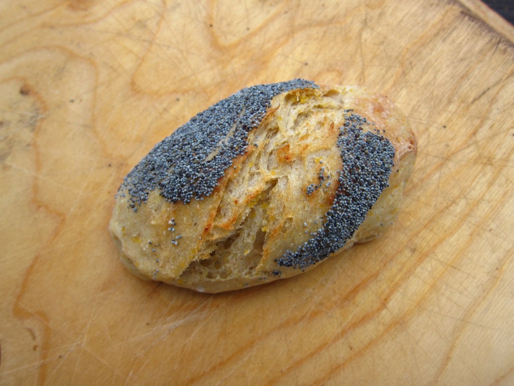 Ein frisch gebackenes Brötchen mit Mohnsamen auf der Oberseite, platziert auf einem Holzbrett.