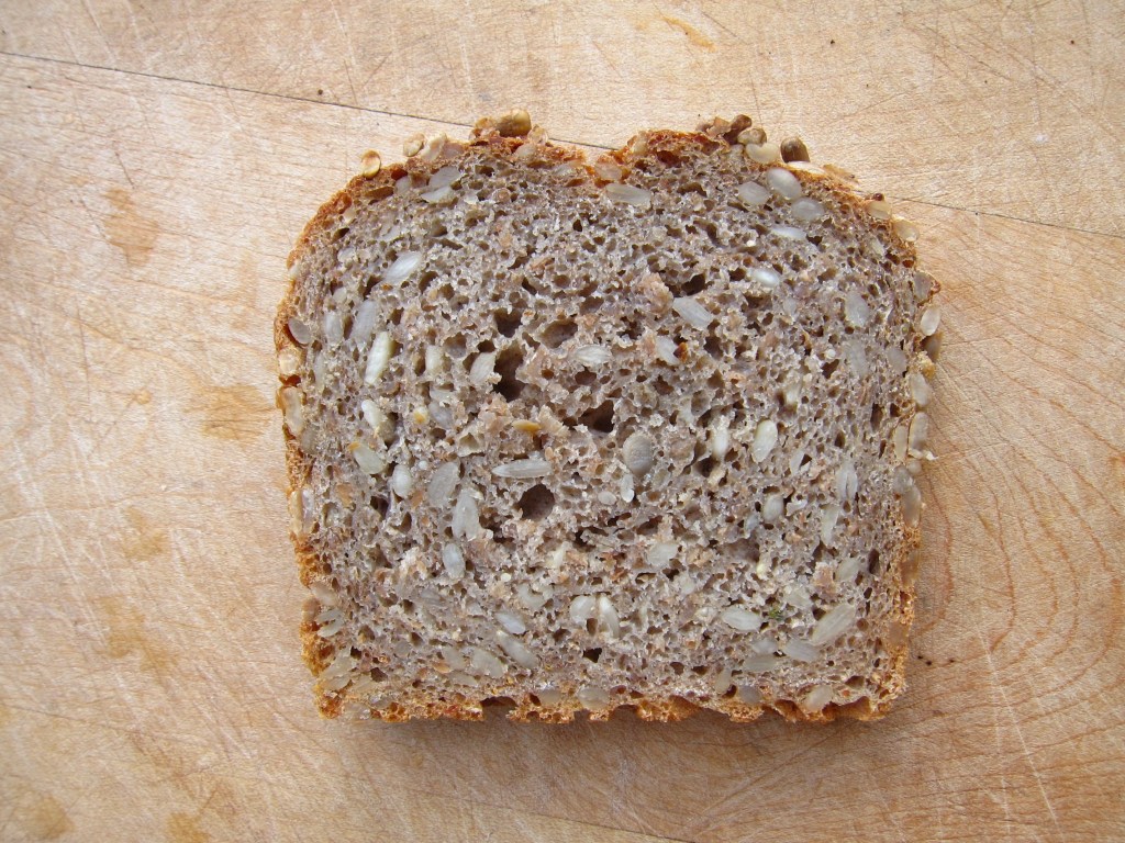Eine Scheibe Vollkornbrot mit sichtbaren Körnern und einer dichten Krume, auf einem Holzbrett.