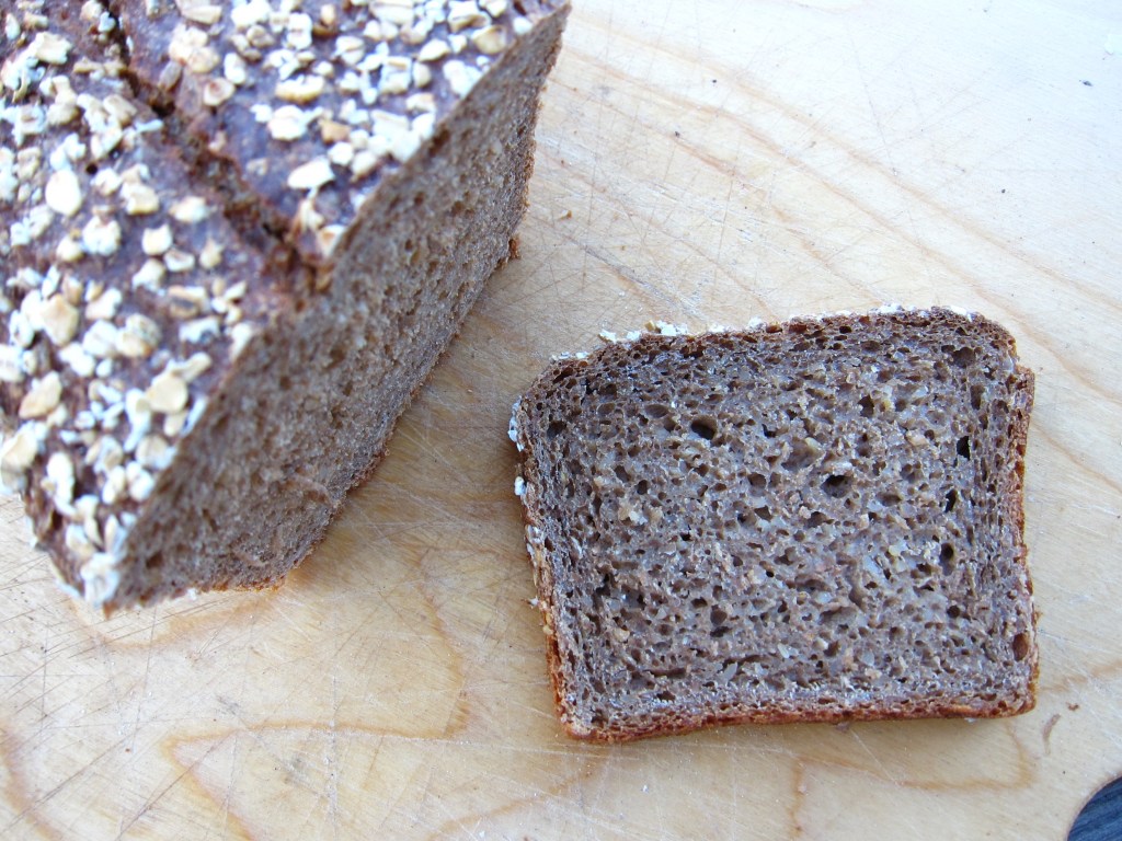 Ein Laib Vollkornbrot mit Haferflocken obendrauf, auf einem Holzbrett, mit einem geschnittenen Stück daneben.