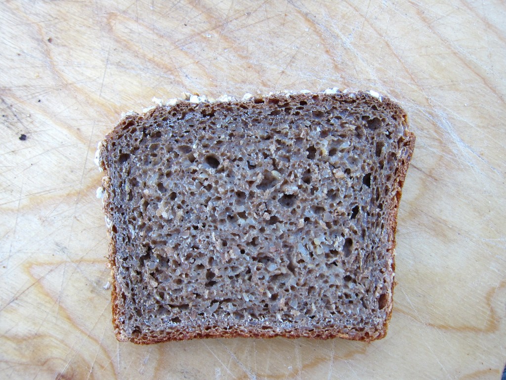 Eine Scheibe dunkles Brot auf einem Holzbrett mit sichtbarer Porung und Krume.