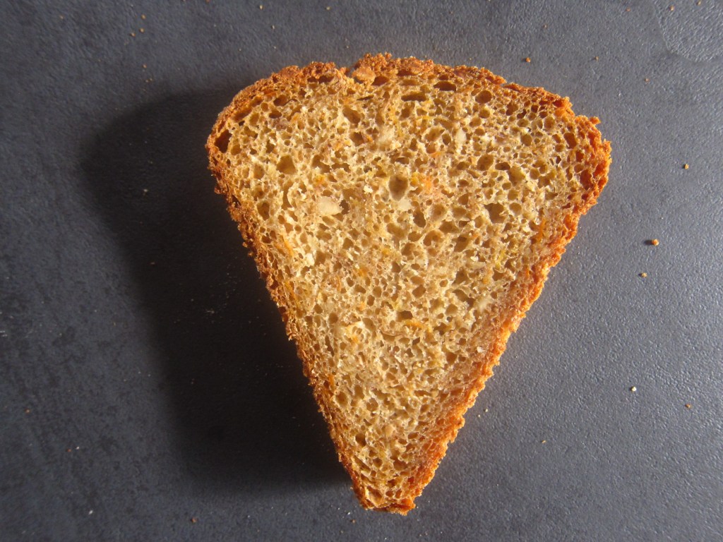 Eine Scheibe rustikales Brot mit einer goldbraunen Kruste und einer groben, luftigen Innenstruktur auf einem dunklen Untergrund.