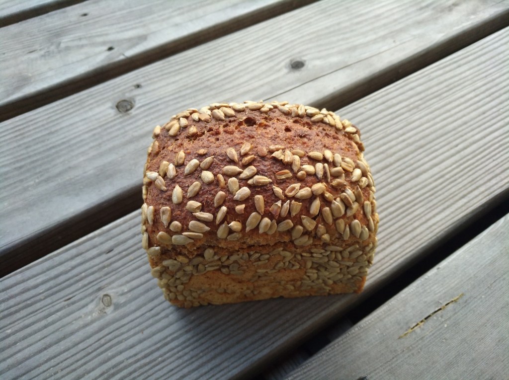 Ein braunes, hochformatiges Brot mit einer knusprigen Kruste, bestreut mit Sonnenblumenkernen und auf einer Holzoberfläche abgebildet.