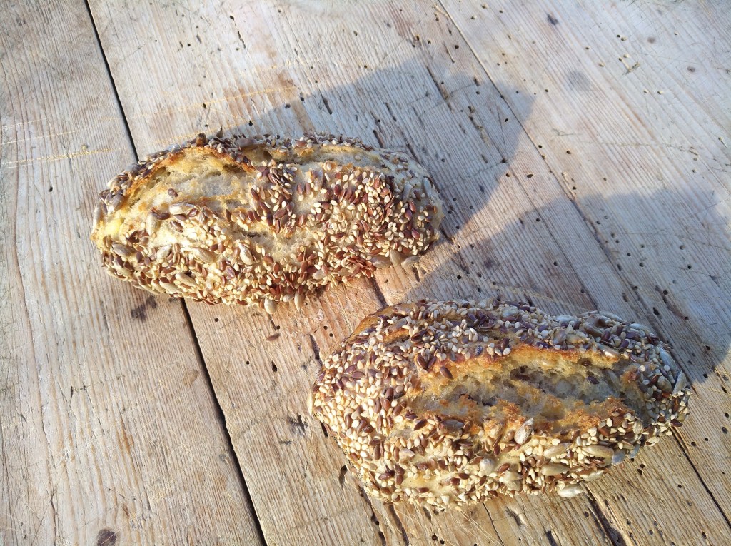 Zwei rustikale Brötchen, mit einer knusprigen Kruste und einer Mischung aus Sonnenblumen- und Leinsamen, auf einem Holzbrett platziert.