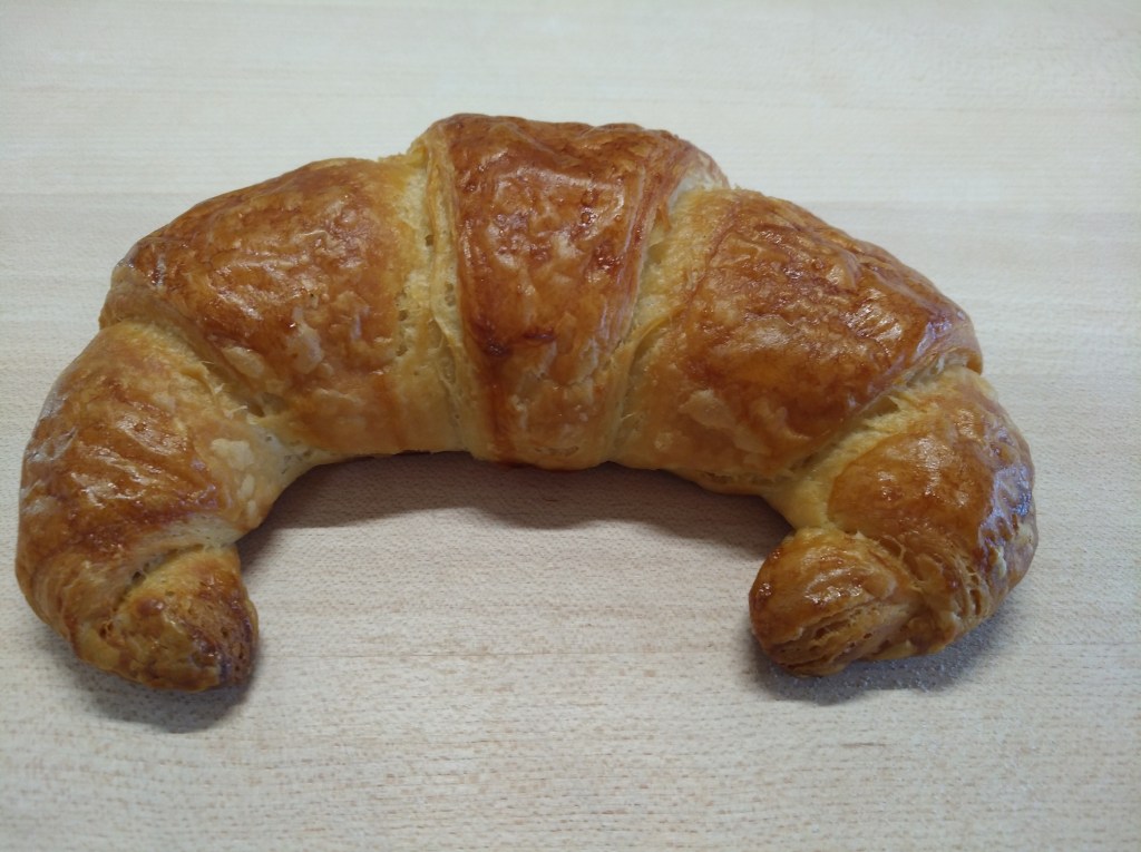 Ein frisch gebackenes Croissant mit goldbrauner, knuspriger Oberfläche, auf einem Holzbrett platziert.