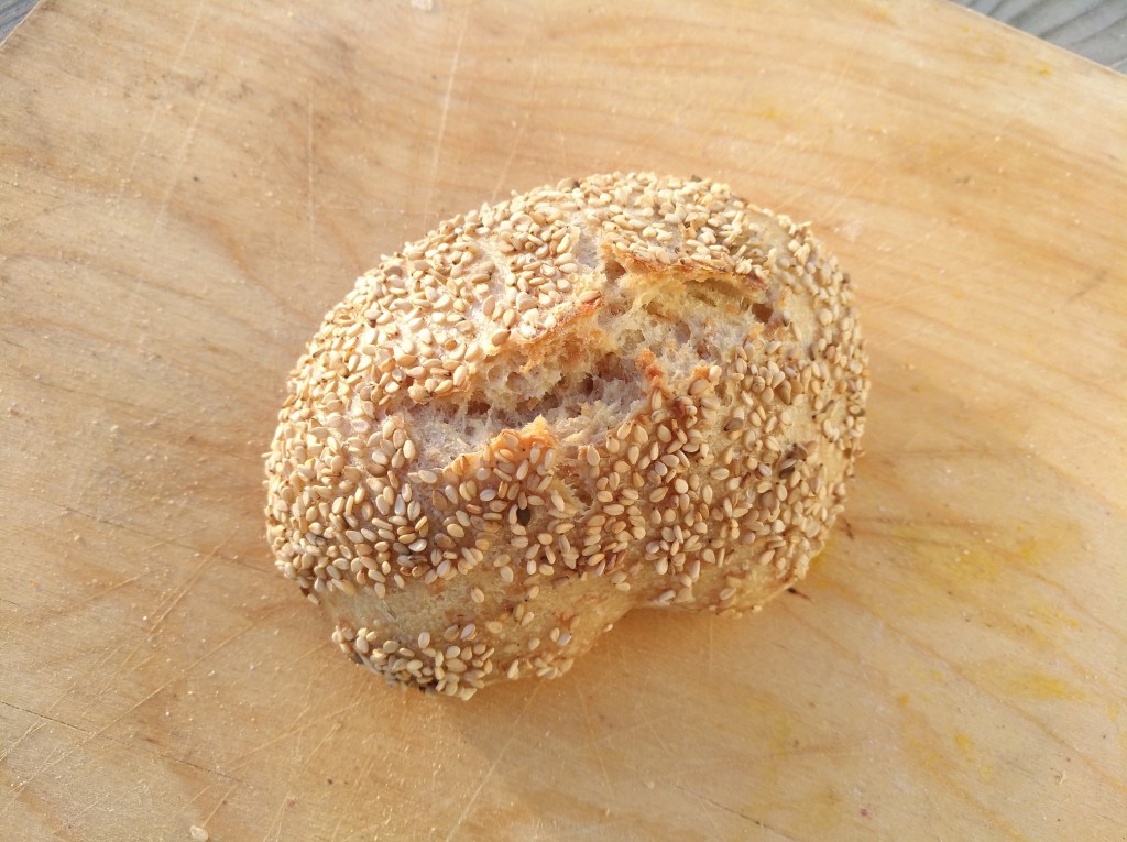 Ein rundes Brötchen mit einer goldbraunen, mit Sesam bestreuten Kruste auf einem Holzbrett.