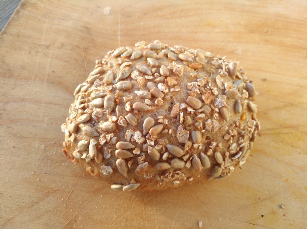 Ein dunkelbraunes Brötchen mit einer knusprigen Kruste, bestreut mit Sonnenblumenkernen und Haferflocken, auf einem Holzbrett platziert.