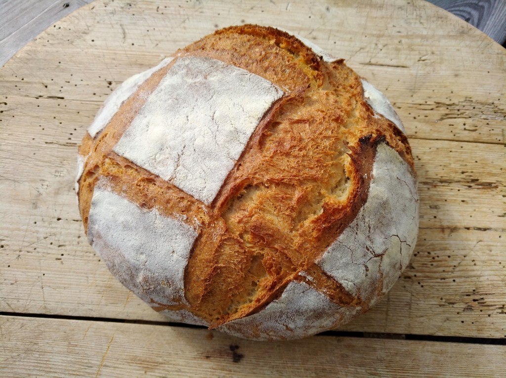 Ein rundes, gebräuntes Brot mit einer knusprigen Kruste und einer leicht mehlbestäubten Oberfläche, auf einem Holzbrett platziert.
