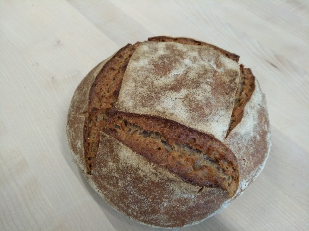 Ein frisch gebackenes, rundes Sauerteigbrot mit einer goldbraunen Kruste und Schnittmuster auf der Oberfläche, auf einem hellen Holzuntergrund.