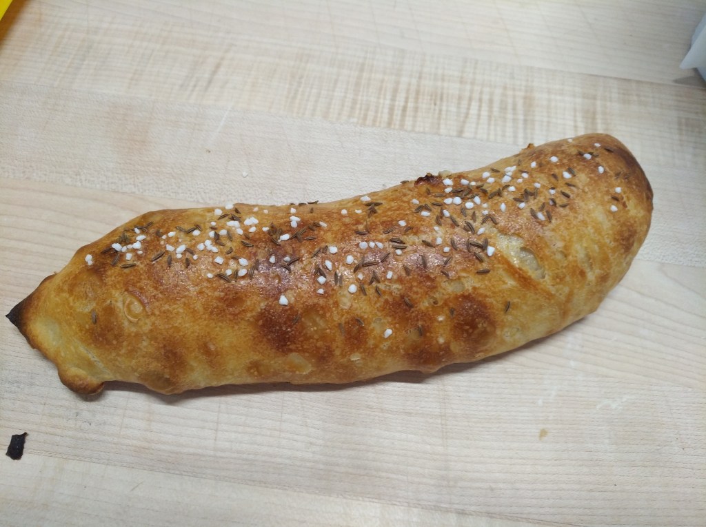 Ein frisch gebackenes Brot mit einer goldbraunen Kruste, bestreut mit Kümmel und grobem Salz, auf einem Holzbrett.