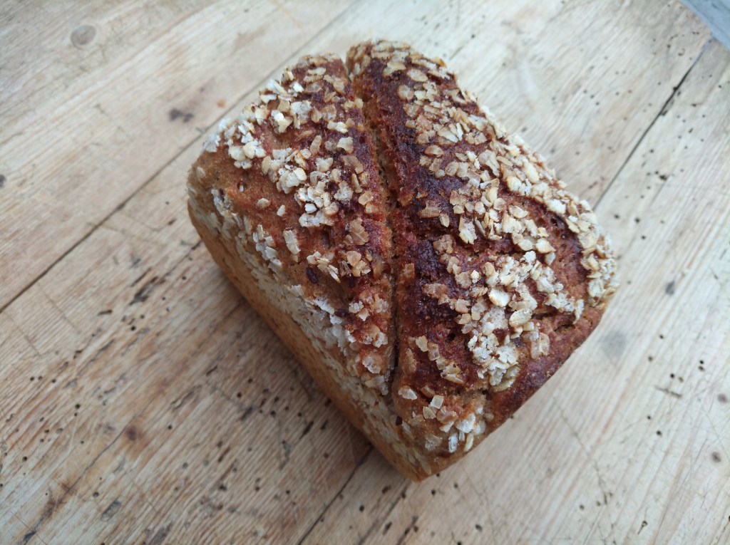 Ein frisch gebackenes Brot mit einer knusprigen Haferkruste und einer goldbraunen Oberfläche auf einem Holzuntergrund.