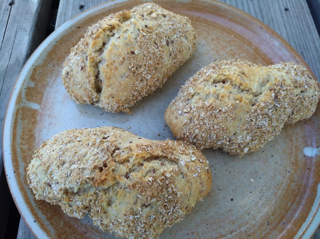 Drei goldene Brötchen mit einer knusprigen Haferflockenoberfläche, angerichtet auf einem rustikalen Keramikteller.