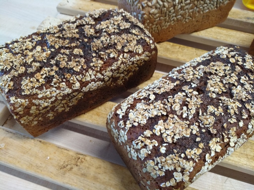 Zwei frisch gebackene Brote mit einer Haube aus Haferflocken, auf einer Holzunterlage platziert.