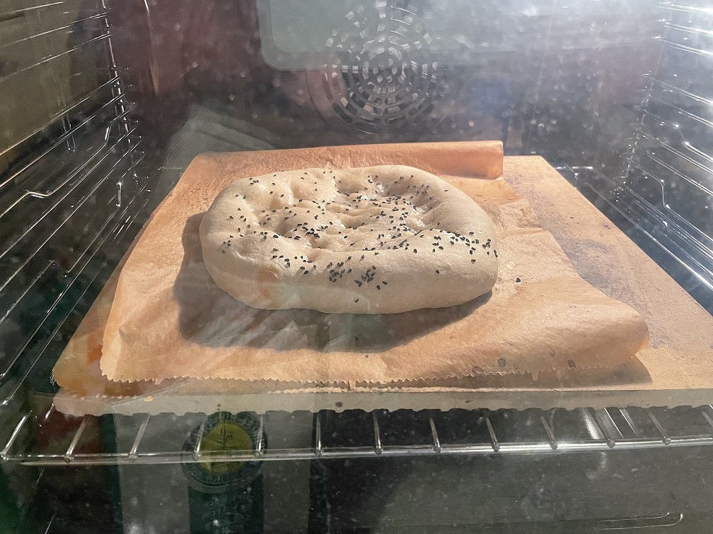 Ein Fladenbrot, das im Ofen backt, auf einem Backpapier platziert, mit Schwarzkümmel bestreut.