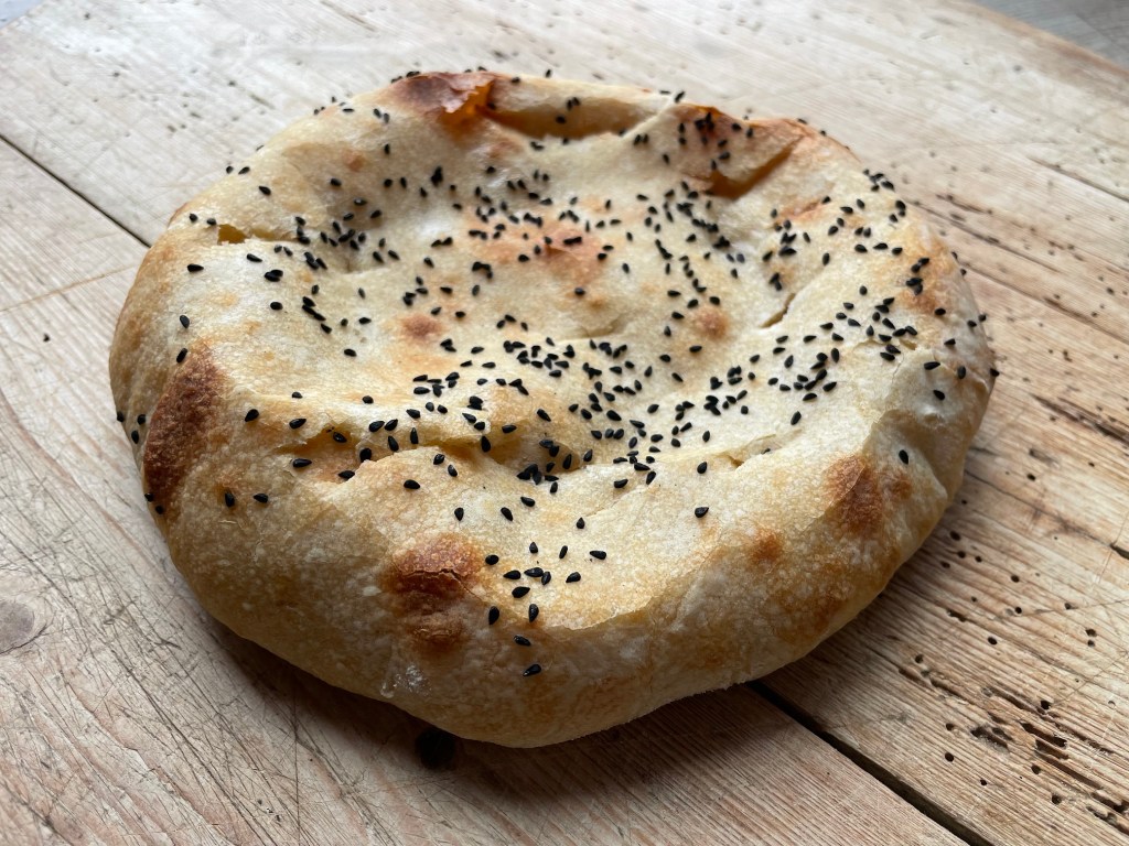 Ein frisch gebackenes Fladenbrot mit schwarzem Kümmel auf einem Holzbrett.