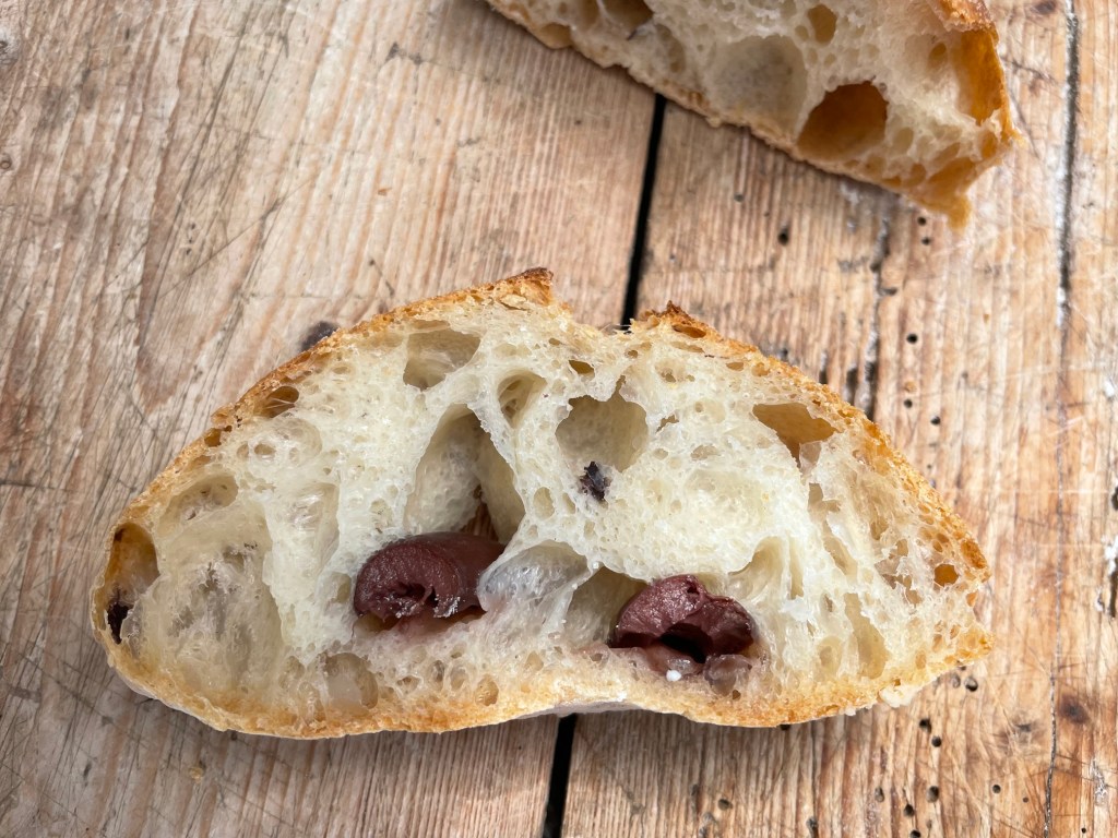 Aufgeschnittenes Brot mit einer lockeren Krume und sichtbaren Olivenstücken, auf einem Holztisch präsentiert.