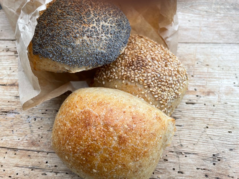 Drei Brötchen mit verschiedenen Toppings, darunter Mohn und Sesam, in einer Papiertüte auf einer Holzoberfläche.